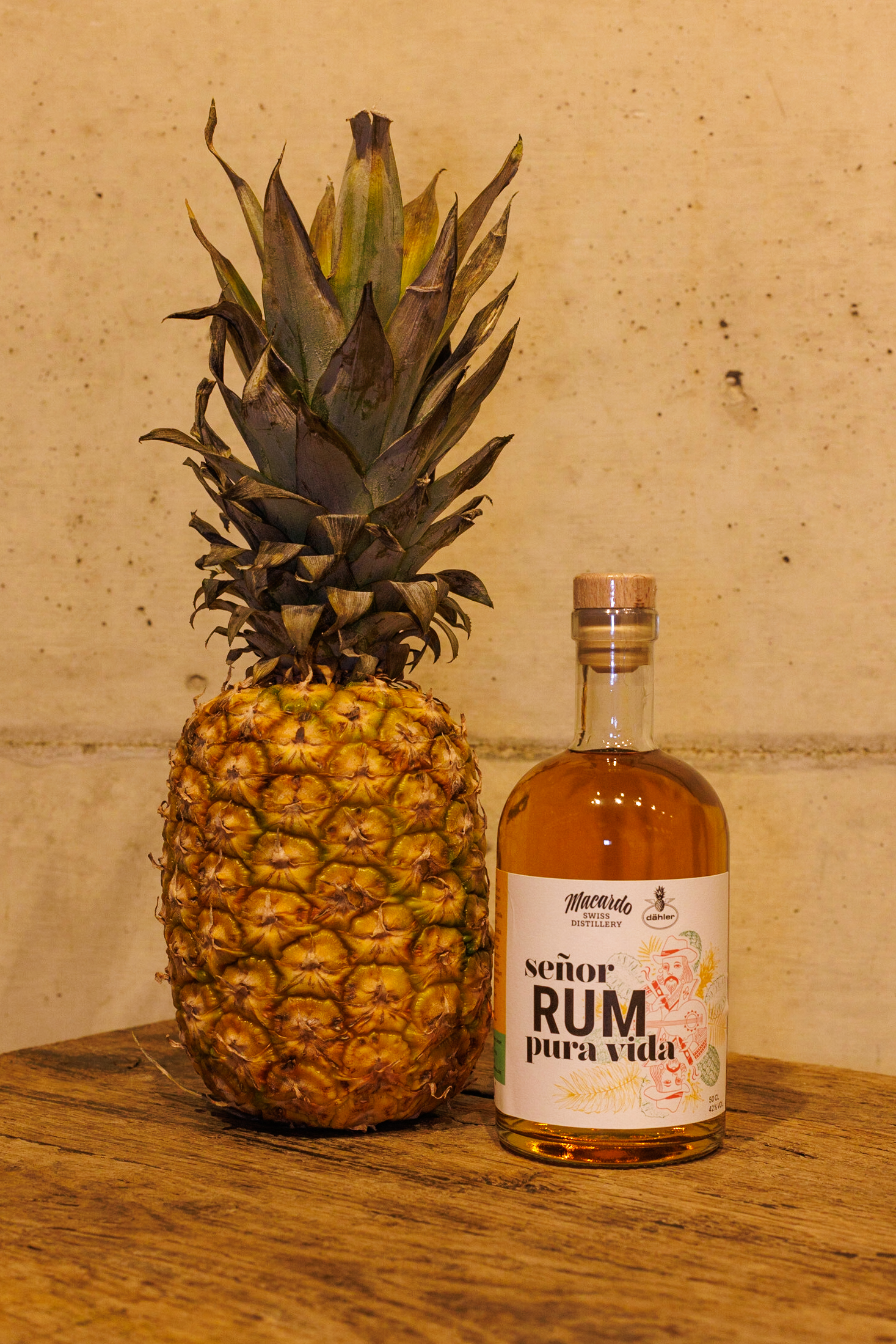 Eine braune Rum Flasche der Marke "Señor Rum - Pura Vida" steht auf einem Holztisch, direkt daneben eine reife Ananas mit grünen Blättern. Die Flasche hat ein weißes Etikett mit bunten Verzierungen.